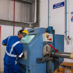 "Technician wearing safety gear operating a blasting or testing machine for surface treatment or inspection of a cylindrical metal component in an industrial setting."