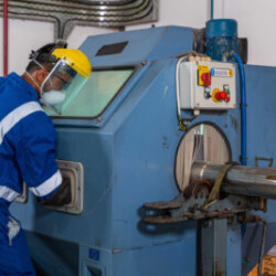 Technician wearing safety gear operating a blasting or testing machine for surface treatment or inspection of a cylindrical metal component in an industrial setting.