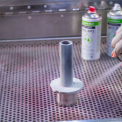 "Technician applying a white developer spray during liquid penetrant testing (LPT) on a cylindrical metal component, ensuring surface defects become visible."